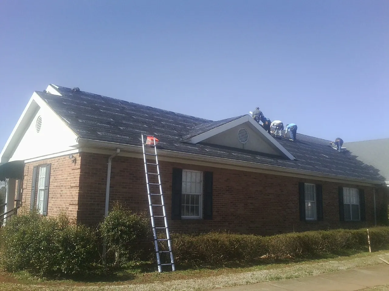 Crew tearing shingles off a brick ranch roof with a ladder set — Metal Roof Repair work by our crew in Hurley