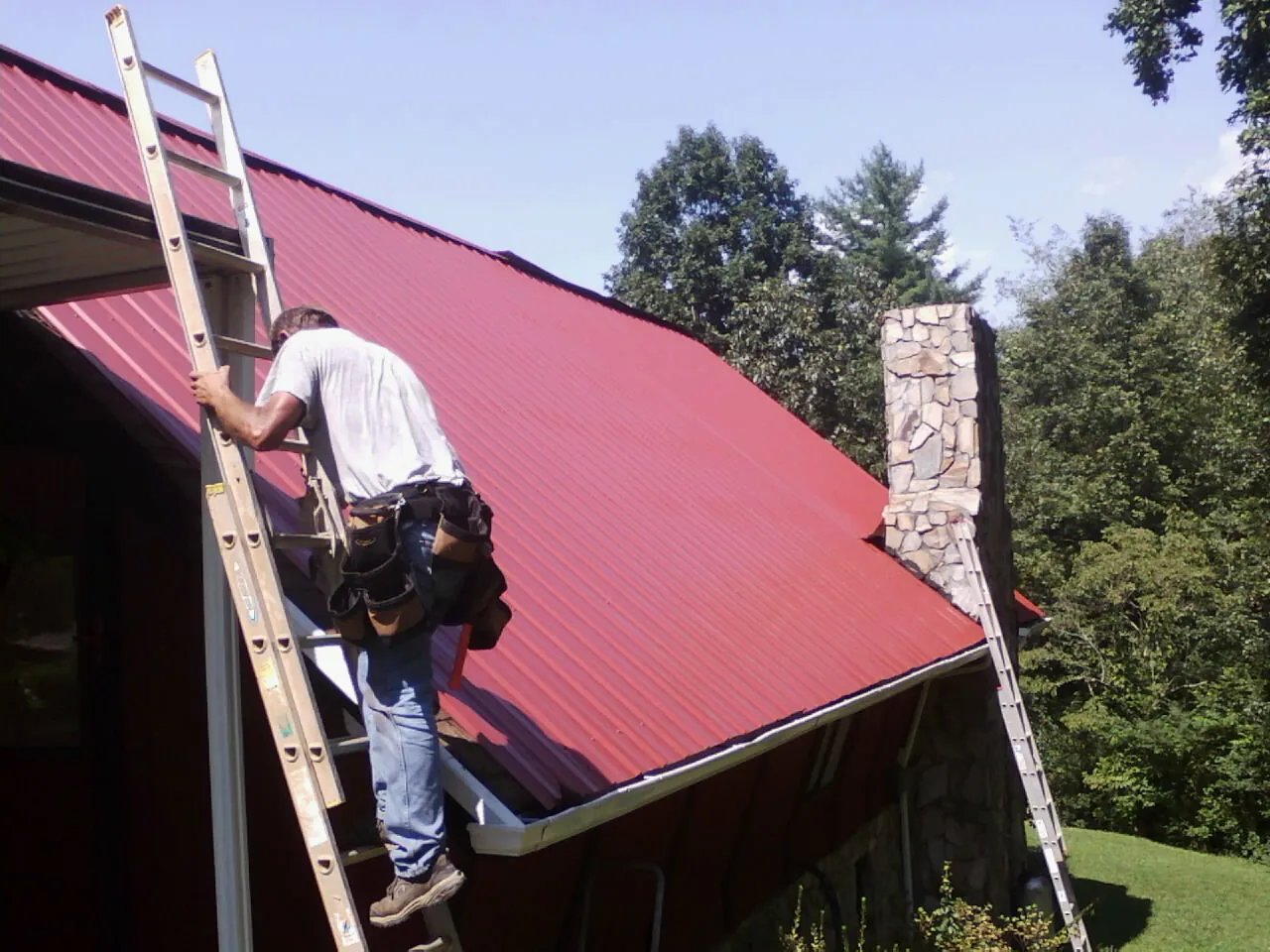 Roofer on a ladder beside a red metal roof and stone chimney — Soffit & Fascia Repair by Roofing Brothers in Hurley